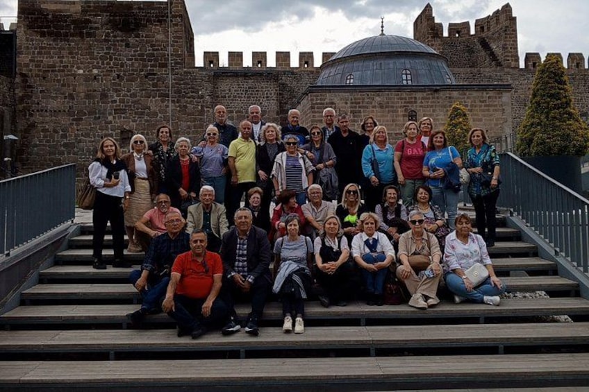 A group of people posing in front of a historic stone building with a dome and a castle-like structure in the background. 1