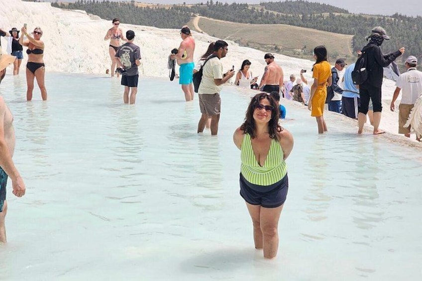 A group of people in a shallow, light blue water body, likely a natural pool or hot spring, with a mountainous landscape in the background. 1