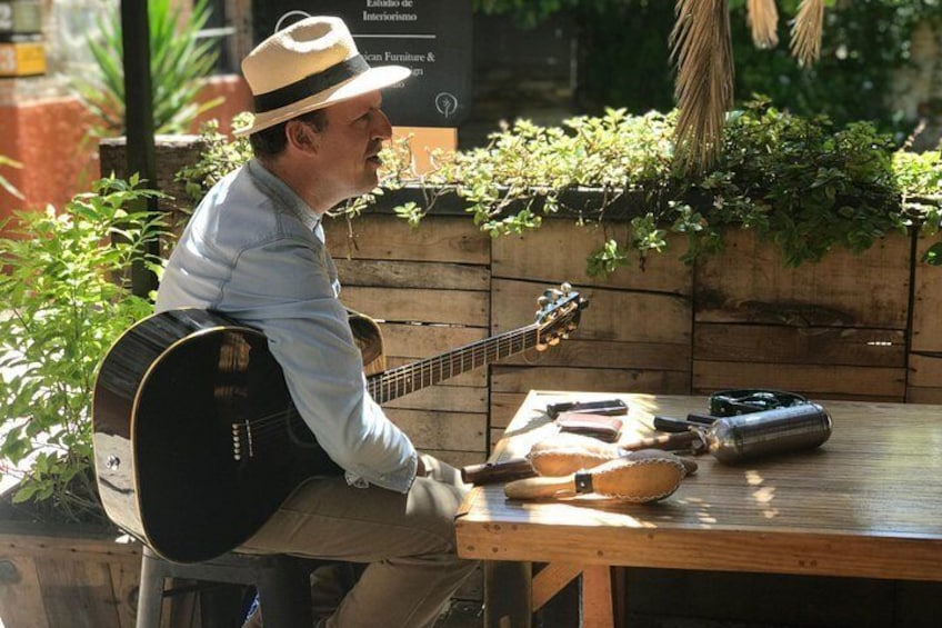 Una persona tocando la guitarra en una mesa de madera, con plantas y un estuche para guitarra cerca. 1