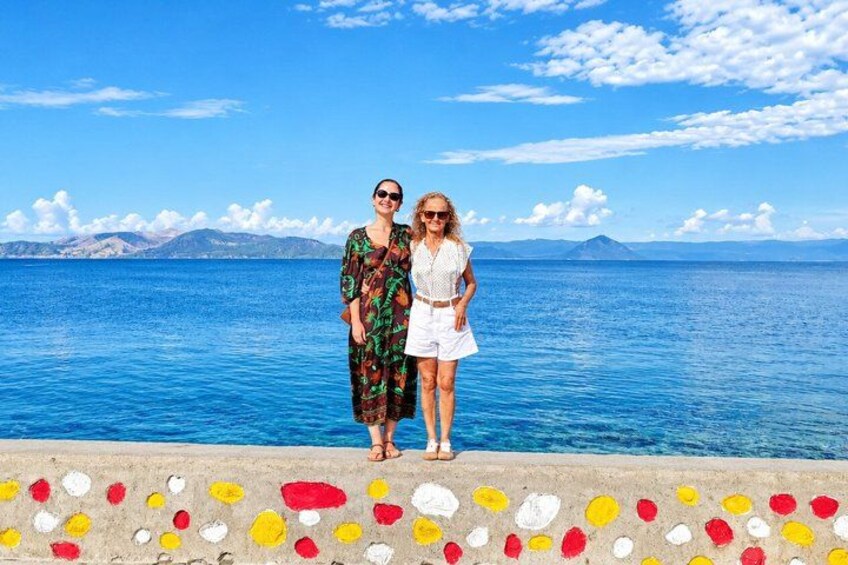 Two people standing on a concrete ledge with a colorful painted border, overlooking a calm sea with mountains in the distance. 5