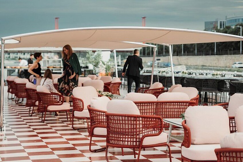 Een overdekt terras met rieten stoelen en tafels, een geruit vloertje en zicht op het water en boten op de achtergrond. 2
