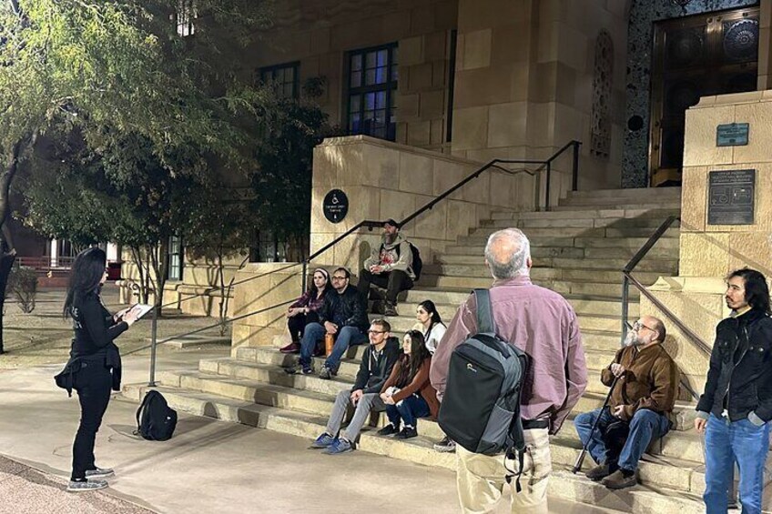 Un grupo de personas sentadas en escalones en un espacio al aire libre, con un edificio de fondo. 1