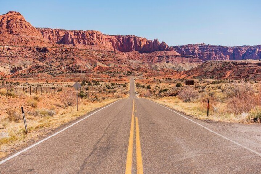 A straight road with double yellow lines, flanked by red rock formations and sparse vegetation. 1