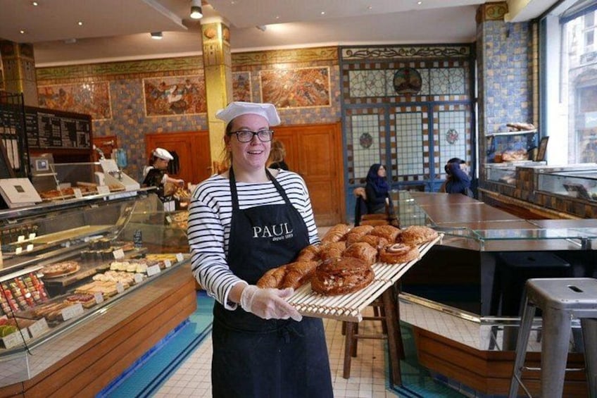 Het interieur van een bakkerij met een persoon die gebak vasthoudt, een vitrine vol gebak en een toonbank met een kassa. 3