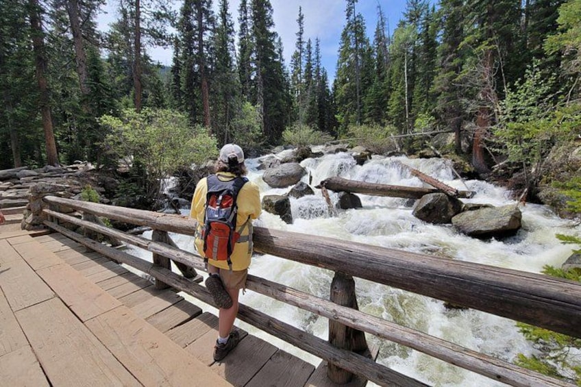 A person standing on a wooden bridge with a backpack, overlooking a fast-flowing river with rocks and trees in the background. 3