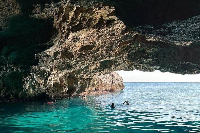 Une falaise rocheuse avec une ouverture menant à une grotte, une eau turquoise et limpide, et deux personnes qui nagent. 1