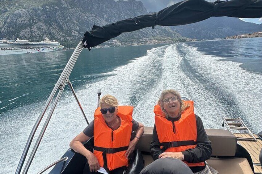 Twee mensen in een boot, met reddingsvesten, met een cruiseschip op de achtergrond en een bergachtig landschap. 3
