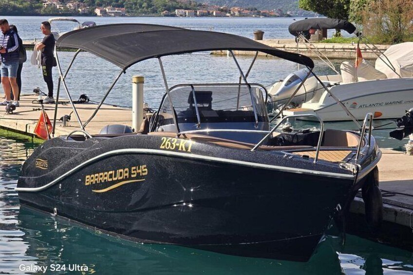 Een zwarte motorboot genaamd 'BARRACUDA 545' ligt afgemeerd in een jachthaven, met mensen en andere boten op de achtergrond. 2