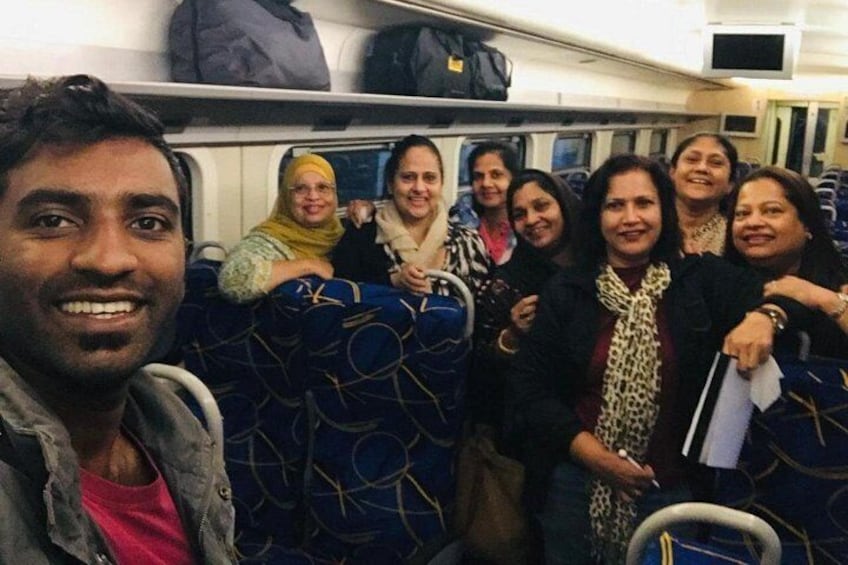 A group of people on a train, some sitting and some standing, with luggage and overhead compartments visible. 4