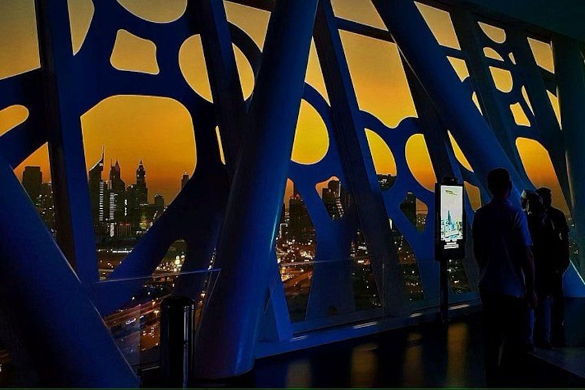 A cityscape at dusk with illuminated buildings, a bridge structure, and people walking. 2