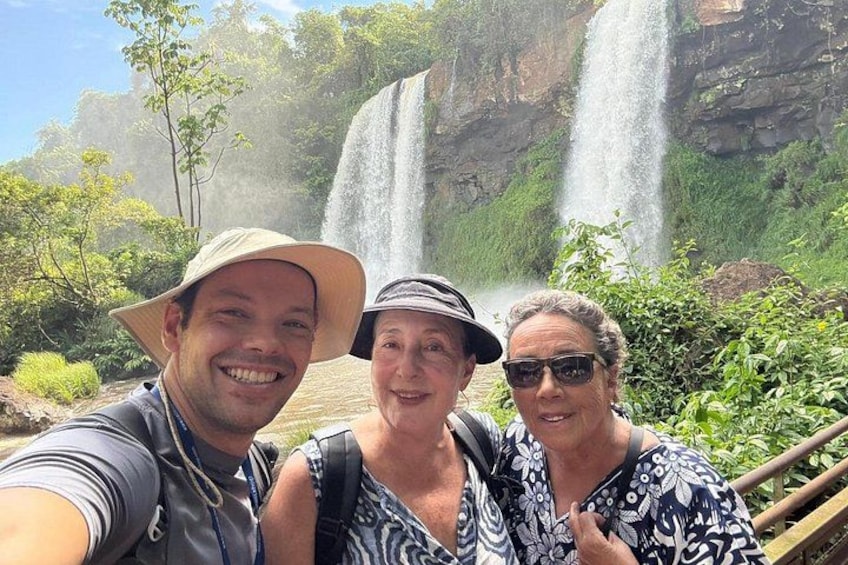 Three people taking a selfie in front of a waterfall with lush greenery and rocky cliffs in the background. 5