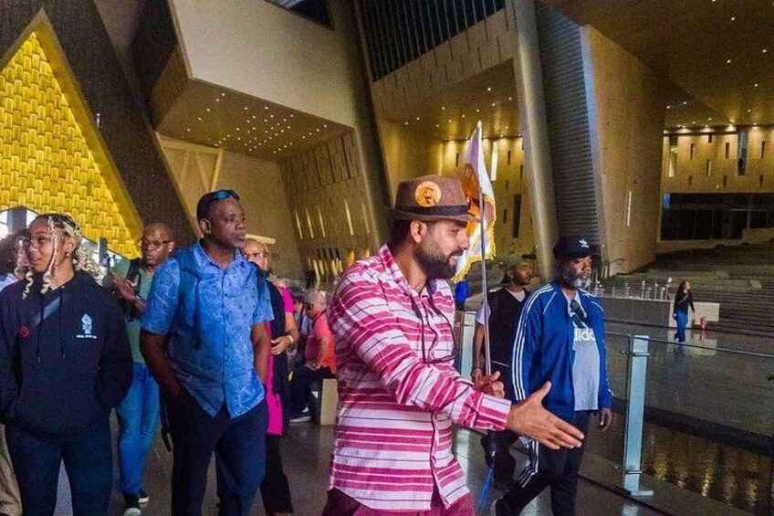 A man in a striped shirt and hat gesturing towards a crowd in a modern building with geometric architecture. 4