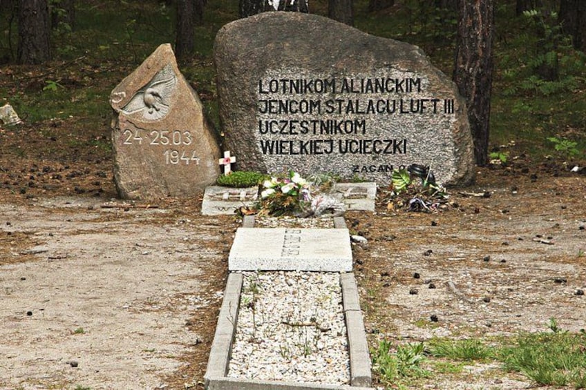 A gravestone with inscriptions and a bird symbol, surrounded by a simple grave marker and some flowers. 1