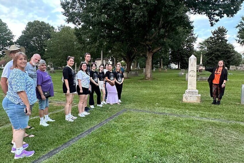 En gruppe mennesker står på en kirkegård med gravsten og et monument i baggrunden. 2