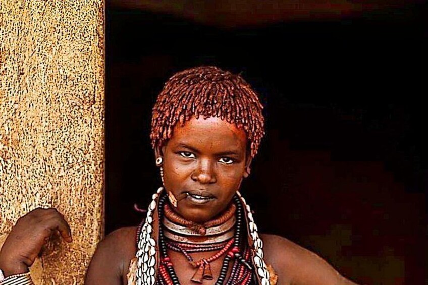 A person with braided hair, wearing traditional jewelry and a patterned garment, standing in front of a textured wall. 4