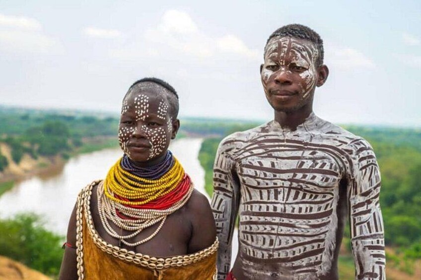 Two individuals standing with a river and greenery in the background. 2