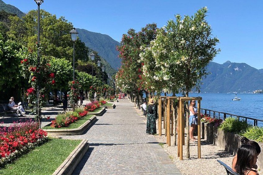 A paved walkway lined with flower beds and trees, leading to a body of water with mountains in the background. 2