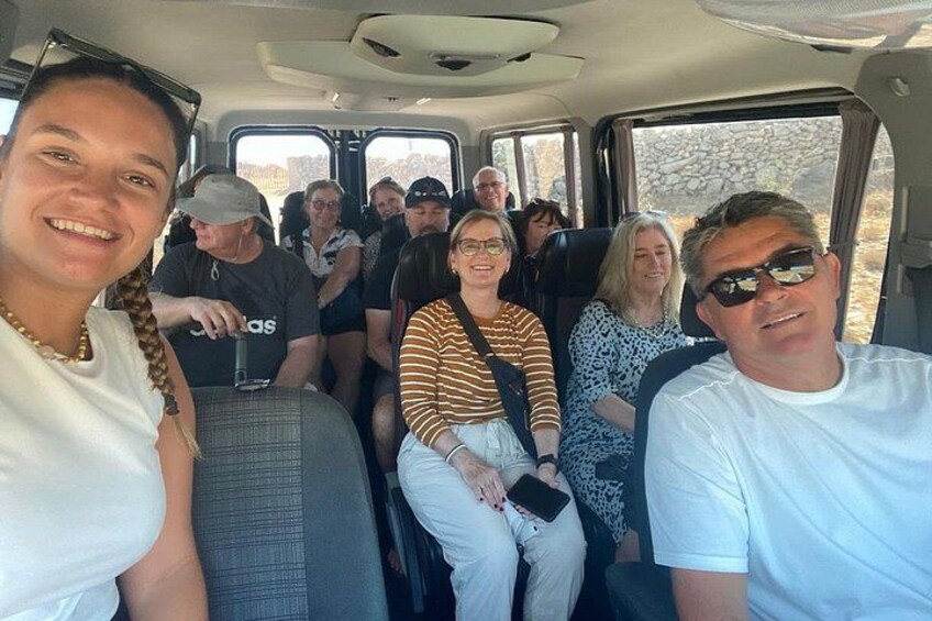 A group of people seated inside a vehicle, likely a tour bus, with windows showing an outdoor landscape. 2
