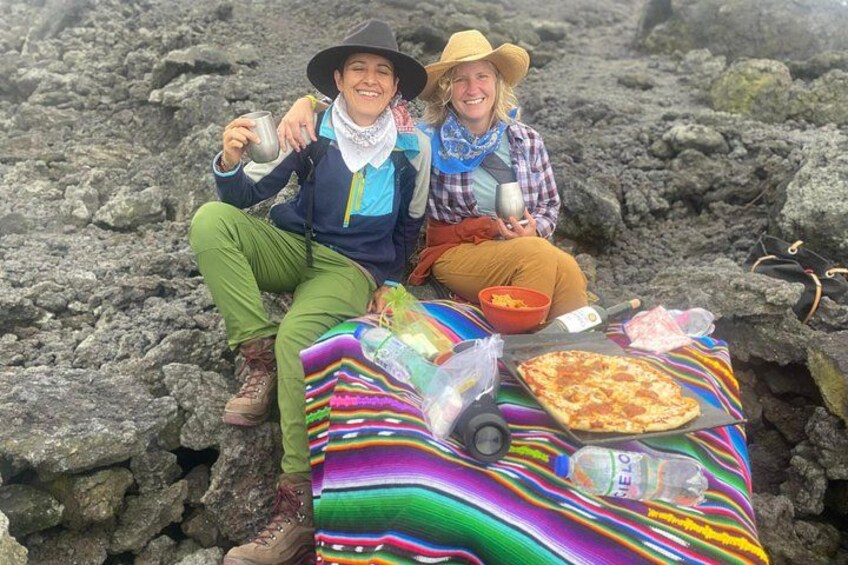 Two people sitting on a colorful blanket with a pizza and a bottle of water on a rocky surface. 2