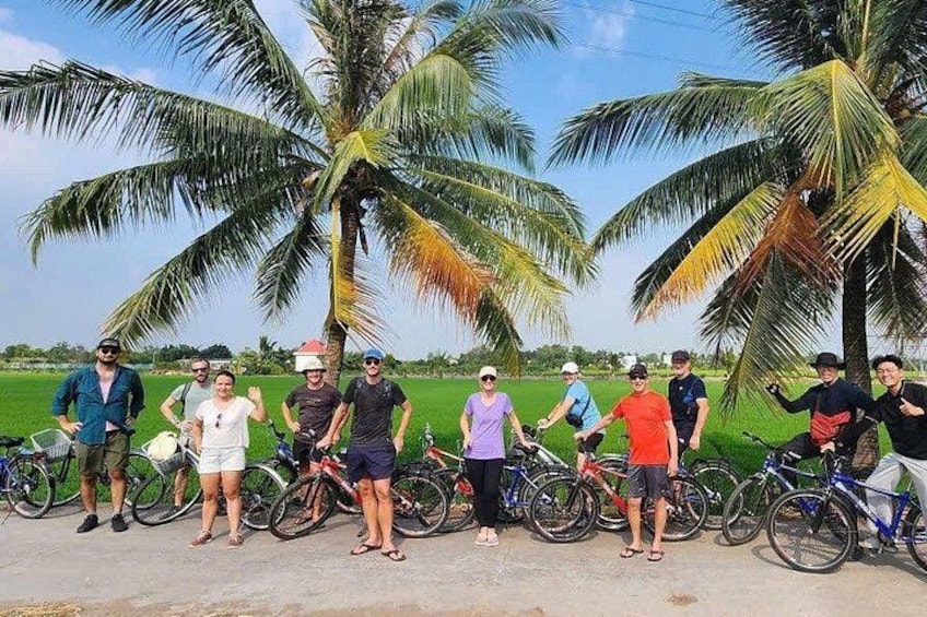 A group of people standing with bicycles in a rural setting with palm trees and a field in the background. 1