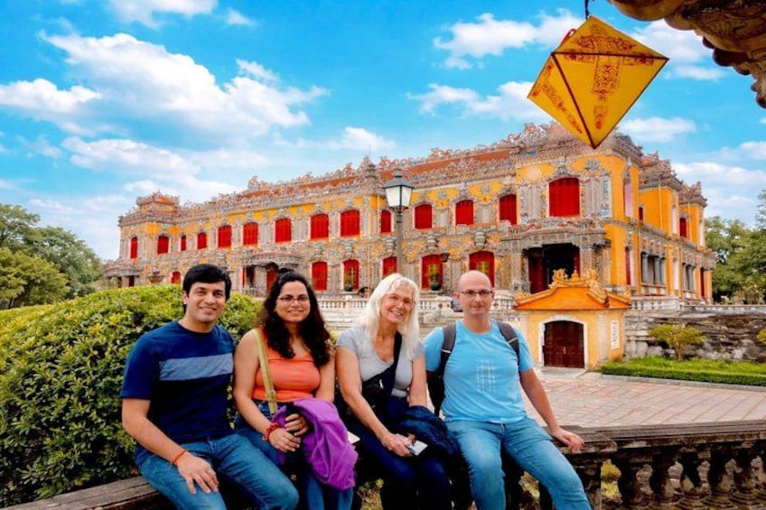 Quatre personnes sont assises sur un banc devant un grand bâtiment orné, avec des fenêtres rouges et un toit jaune. 3