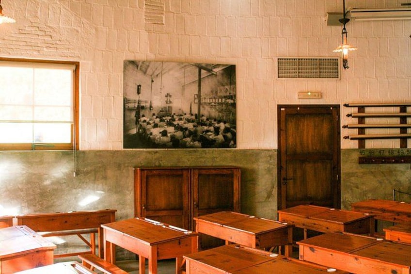 Une salle de classe avec des pupitres et des chaises en bois, une photographie en noir et blanc accrochée au mur et un meuble en bois. 5