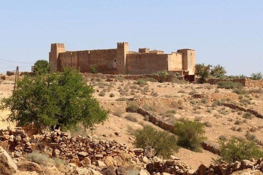A large, ancient stone fortress with multiple towers, surrounded by a dry, hilly landscape with sparse vegetation. 1