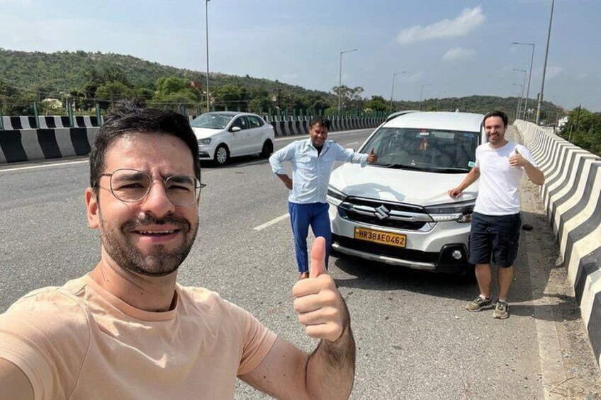 A man giving a thumbs-up, a white car parked on the side of a road, and two people standing behind the car. 3
