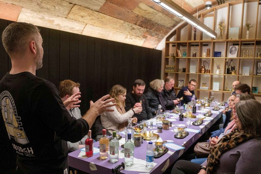 A group of people seated at a long table with various bottles and glasses, in a room with wooden shelves and a stone ceiling. 2