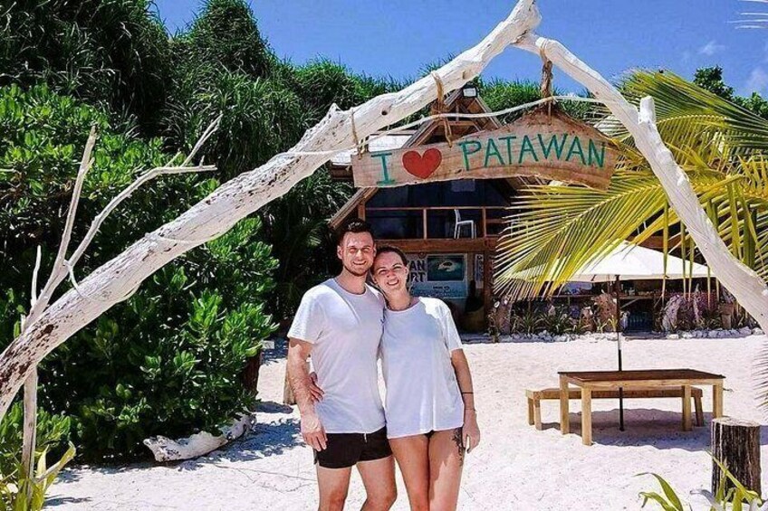 Two people standing under a sign that says 'I ♥ PATAWAN' with a beach and palm trees in the background. 4