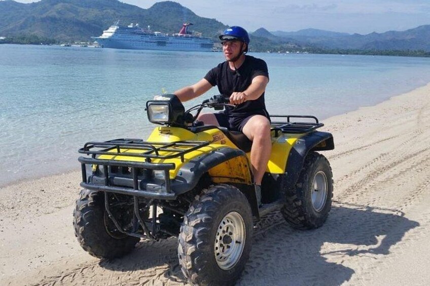A person on a yellow ATV on a sandy beach with a ship in the background. 1
