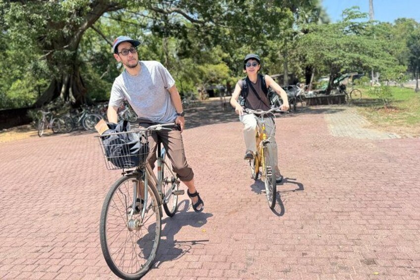 Two people on bicycles in a park with trees and a brick path. 5