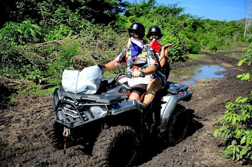 Two people on a quad bike in a muddy area with vegetation around. 5