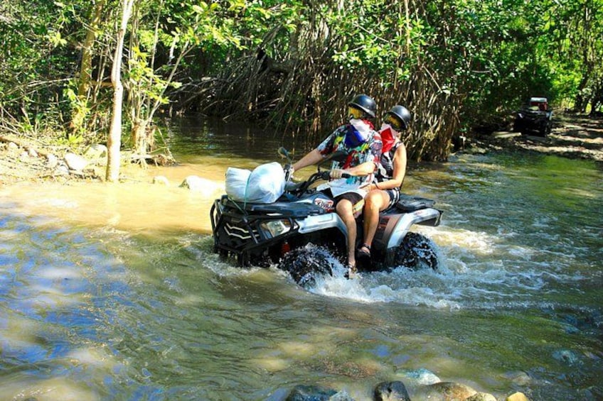 Two people on an ATV navigating through a shallow river surrounded by dense vegetation. 4