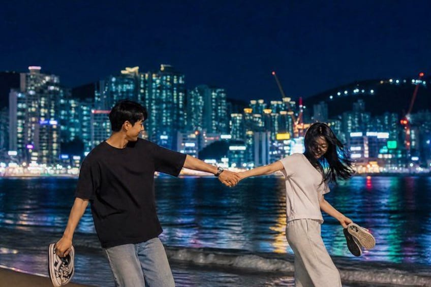 Två personer som håller varandra i handen vid en strandpromenad med en stadsbild i bakgrunden. 5