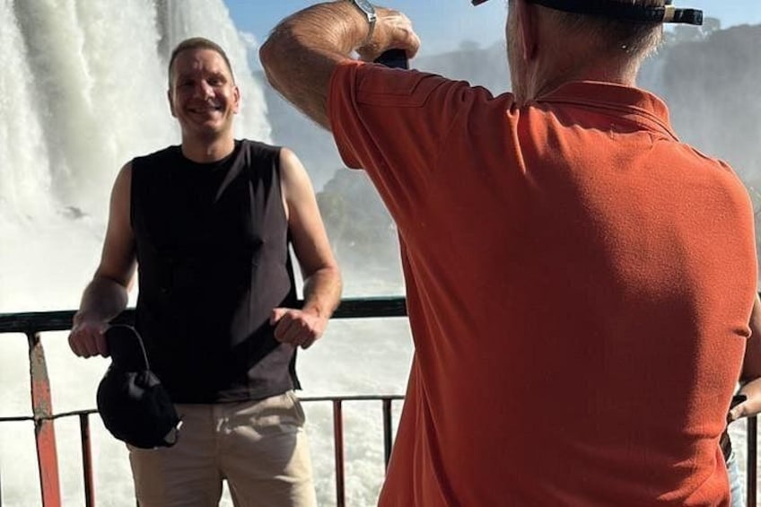 A person in a black sleeveless shirt and beige pants holding a black helmet, standing on a boat with a waterfall in the background. 1