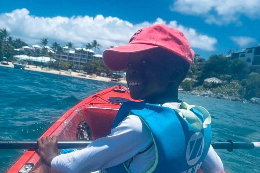 A person in a red kayak on a body of water with a beach and buildings in the background. 2