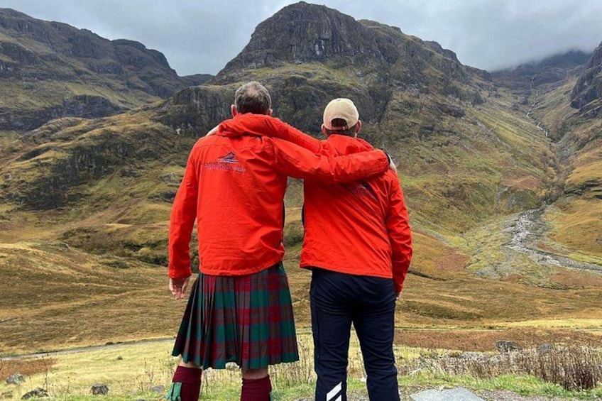 Two people in red jackets and kilts standing on a path, overlooking a mountainous landscape with a river. 3