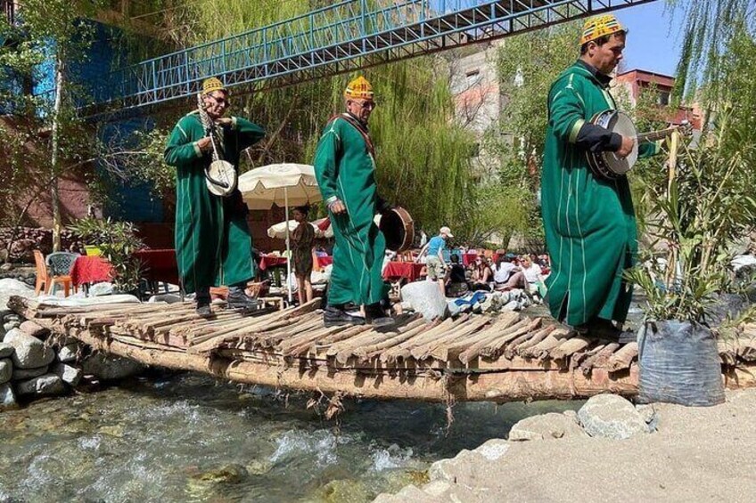 Three individuals in green robes playing traditional stringed instruments on a makeshift wooden bridge over a river. 5