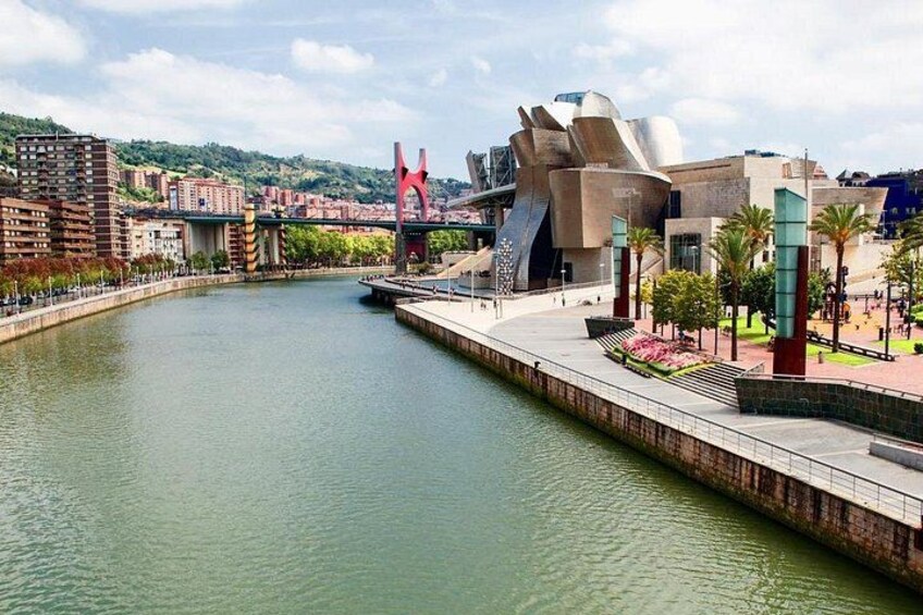 Une rivière traversée par un pont moderne, ornée d’une sculpture remarquable et bordée d’un bâtiment unique à la façade métallique. 3