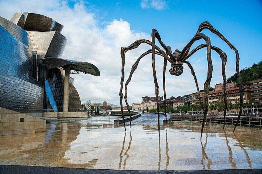 Een modern gebouw met een uniek gebogen ontwerp, een grote metalen spinnenbeeld en een reflecterend wateroppervlak. 1