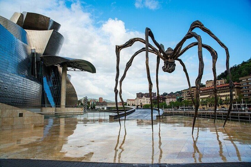Un bâtiment moderne au design original, caractérisé par des courbes, une grande sculpture métallique en forme d’araignée et une surface réfléchissante et humide. 1