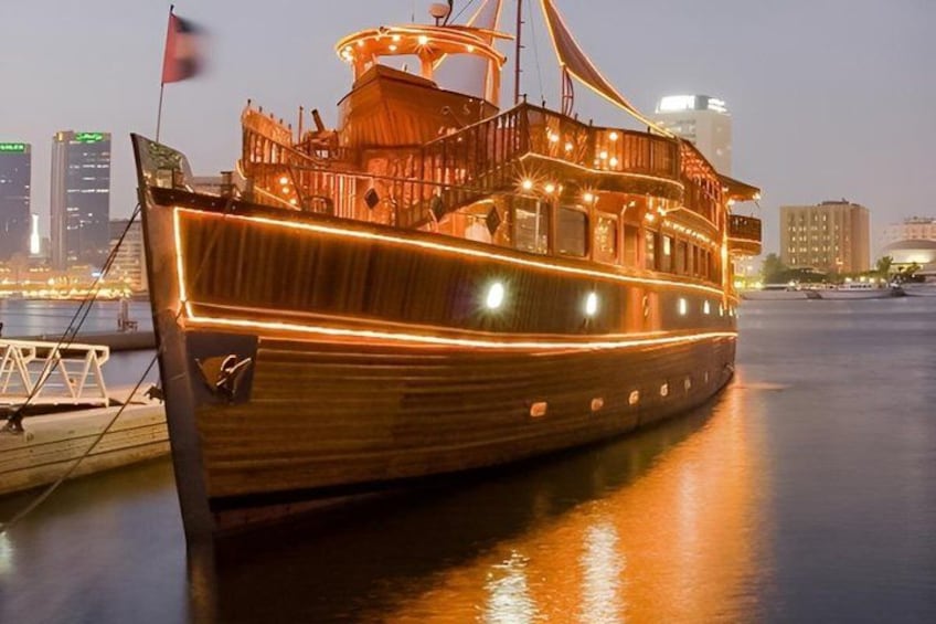 A wooden boat with lights, docked at a marina with city buildings in the background. 1