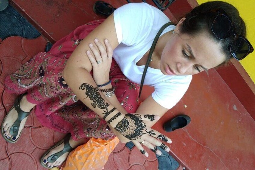 Eine Person mit Henna-Mustern auf Armen und Händen, gekleidet mit einem weißen Oberteil und einem roten Rock, sitzt auf einem Fliesenboden. 5