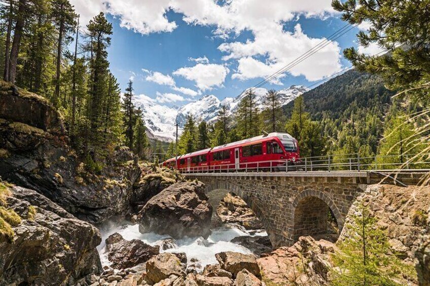 A red train crossing a stone bridge over a rocky stream in a forested mountainous area. 1