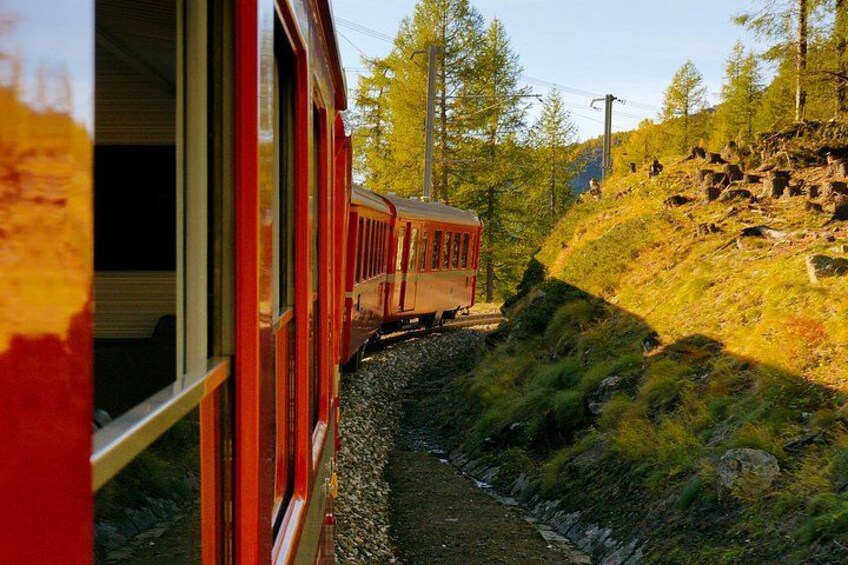 A red train on tracks, surrounded by trees and a rocky hillside. 4