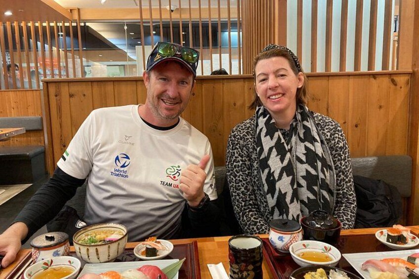 Two people sitting at a table with various dishes, including sushi and bowls of soup, in a restaurant with wooden decor. 4