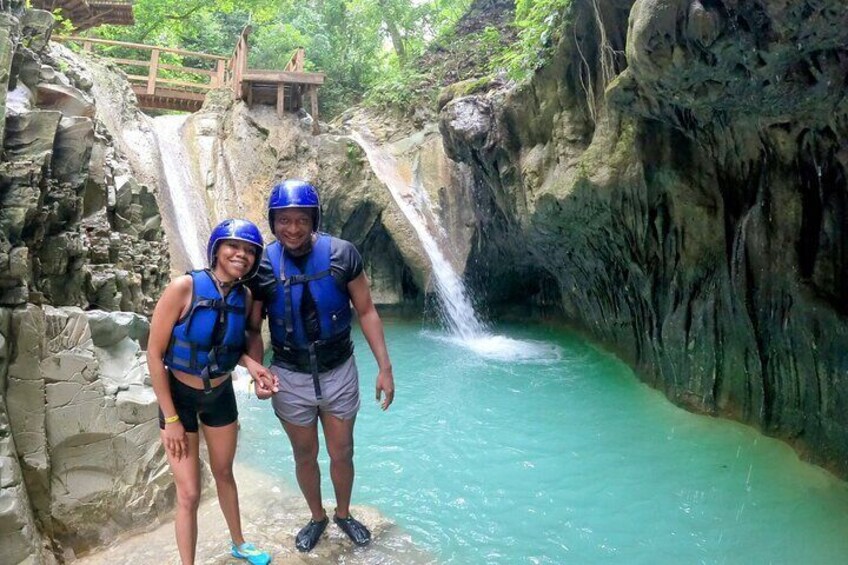Two people in blue life vests standing by a turquoise pool with a waterfall. 3