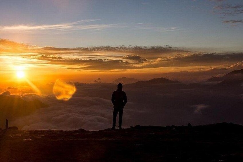 Una persona sobre un pico montañoso durante el amanecer, contemplando un extenso paisaje de nubes y montañas distantes. 3