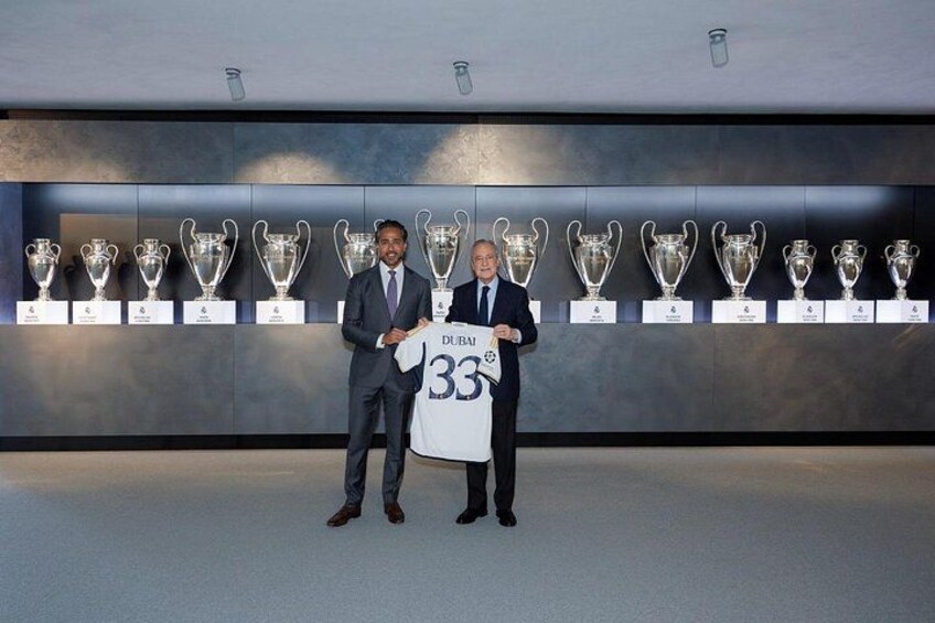 Two men standing in front of a wall displaying trophies. One man holds a jersey with the number 33. 5